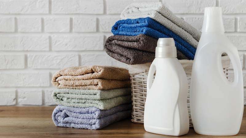 dois frascos de amaciador de roupa brancos ao lado de um cesto de roupa lavada e dobrada; além deste, mais uma pilha de toalhas dobradas em cima da mesa, ao lado do cesto. 